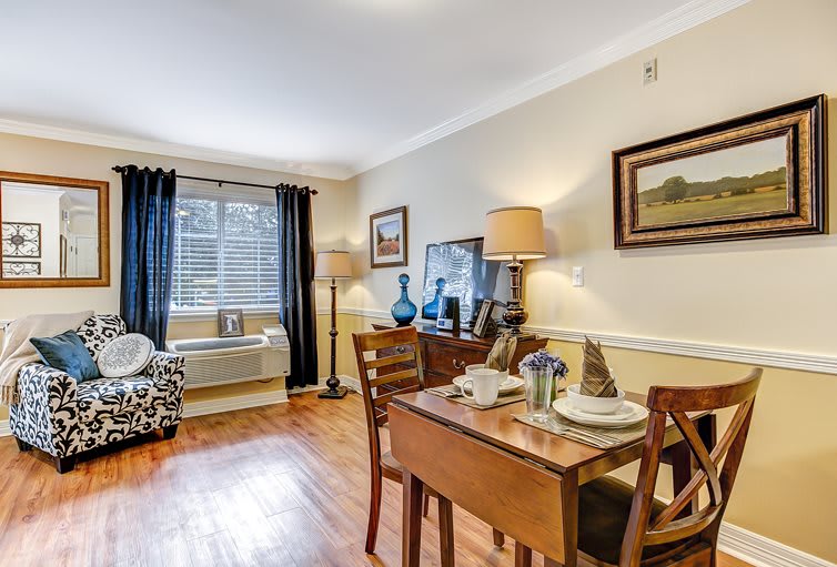 A cozy living and dining space featuring a wooden table with a complete table setting, stylish lamps, and wall art. The room is accented by wood flooring and natural light filtering through the window.