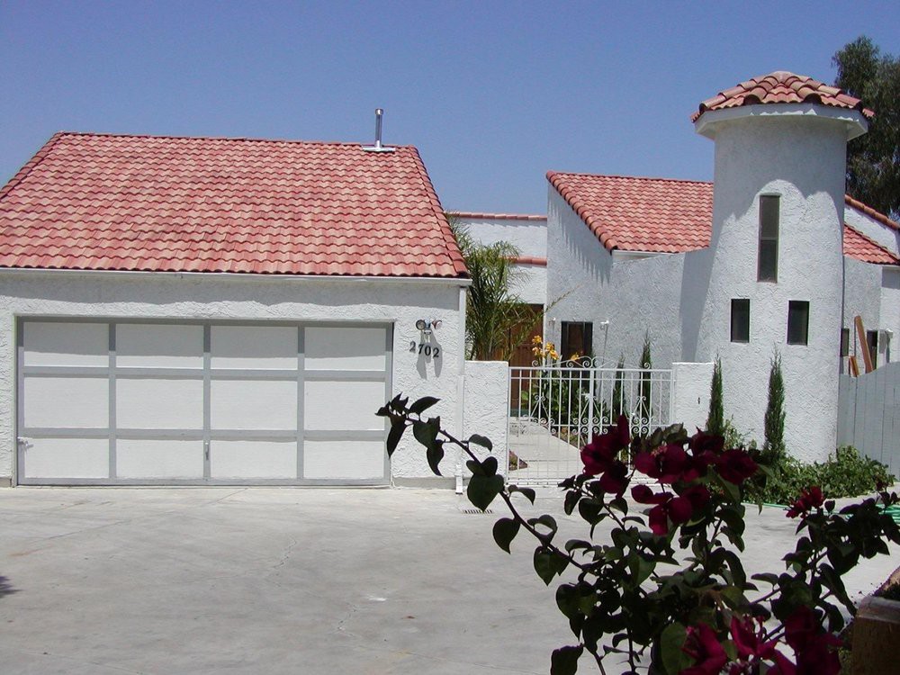 A modern residential house with a tiled roof and attached garage, set against a clear blue sky with flowers in the foreground.