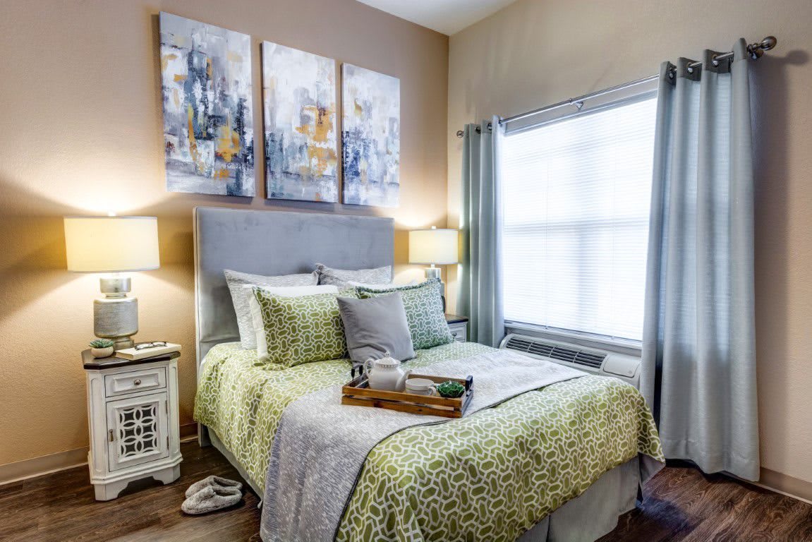 A cozy bedroom with a neatly made bed, adorned with green and gray pillows and bedding. The room features artwork above the headboard, two lamps on nightstands, and a window with curtains.