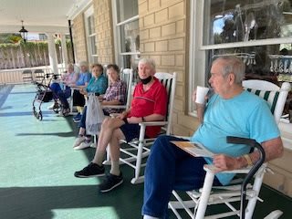 A group of seniors relaxing on a porch, sitting in rocking chairs during the day.