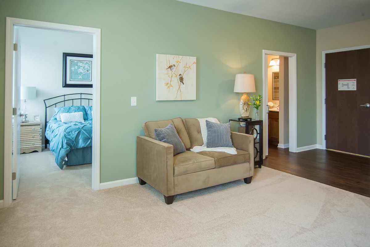 A cozy living area with a beige sofa, green walls, and a view into the bedroom and bathroom.