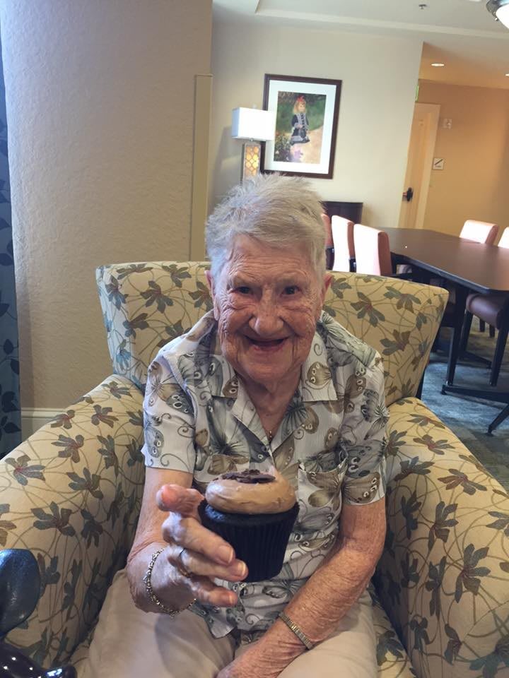 An elderly person smiling while holding a drink, seated comfortably indoors.