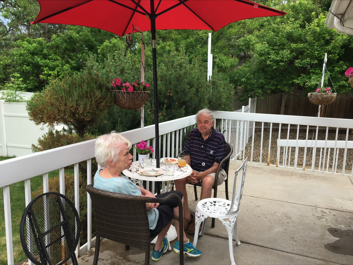 An elderly couple enjoying a conversation on a patio under a red umbrella.