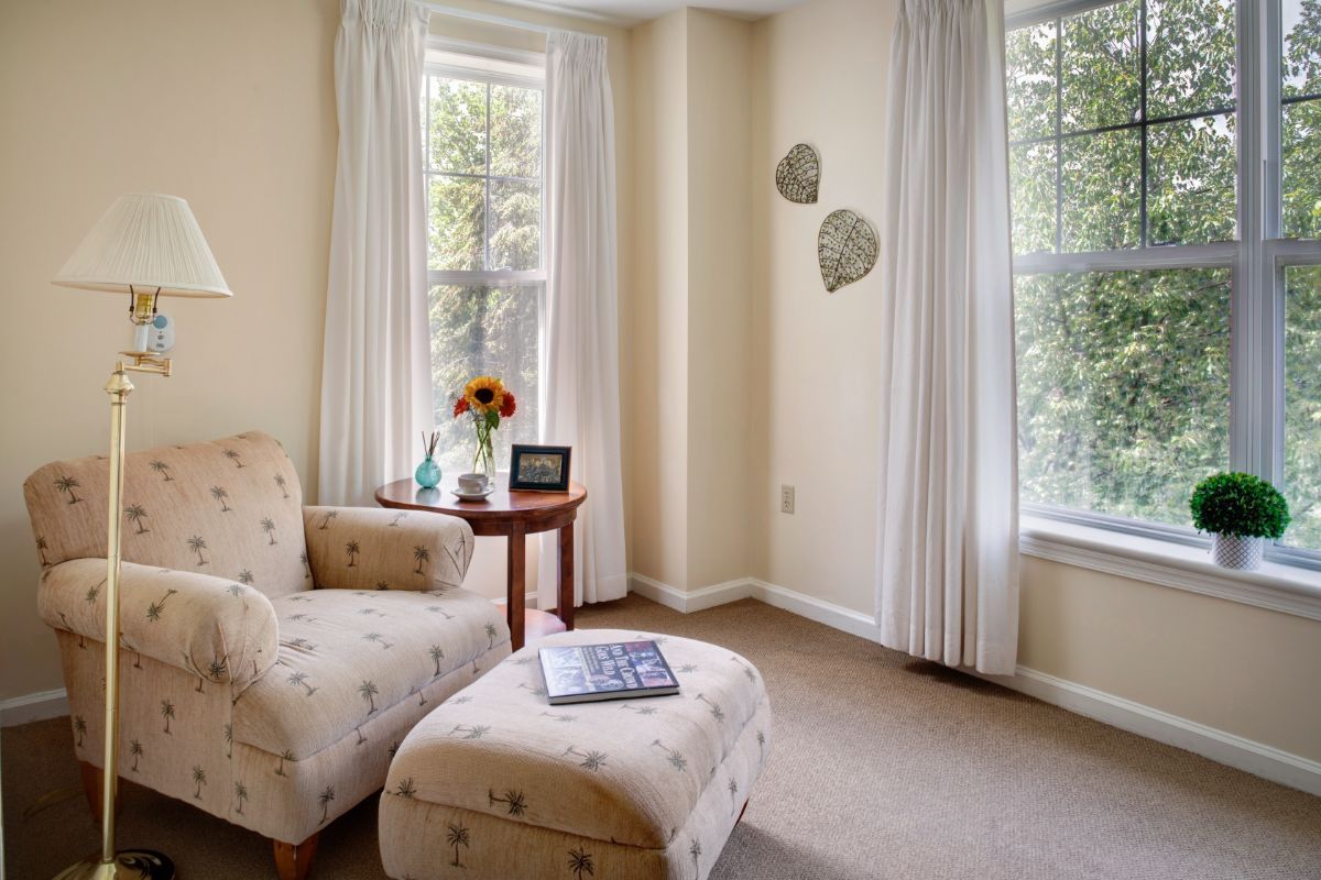 A cozy corner of a living room with a patterned armchair and ottoman, illuminated by natural light from large windows with curtains.