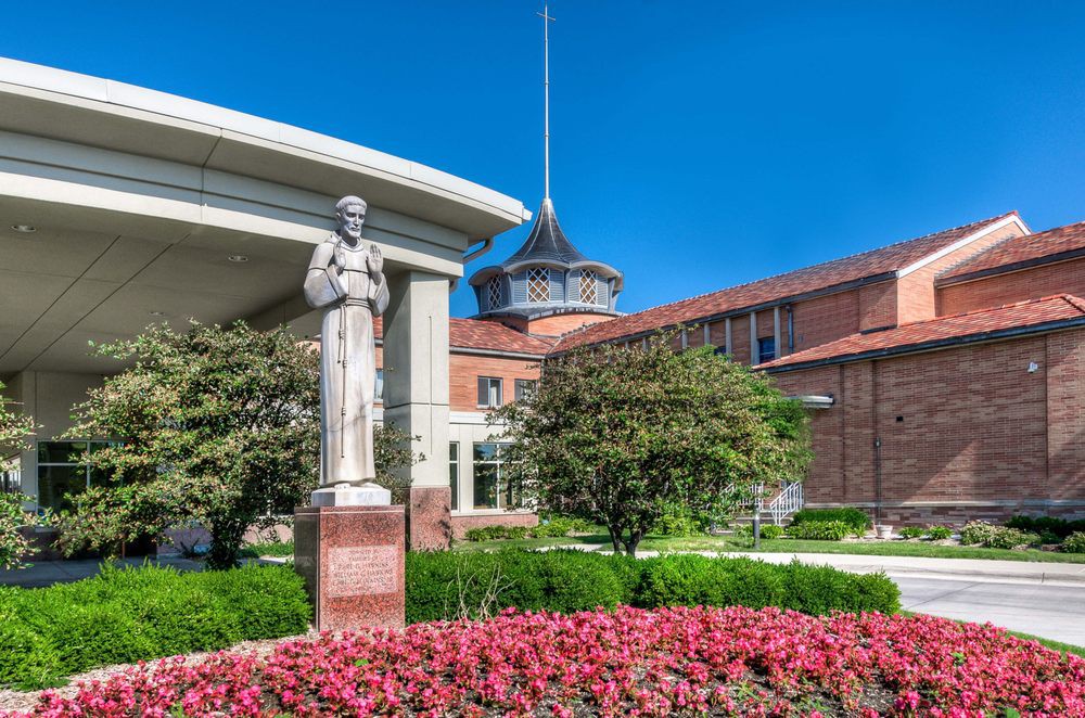 A statue stands in front of a brick building surrounded by vibrant flowers and greenery under a clear blue sky.