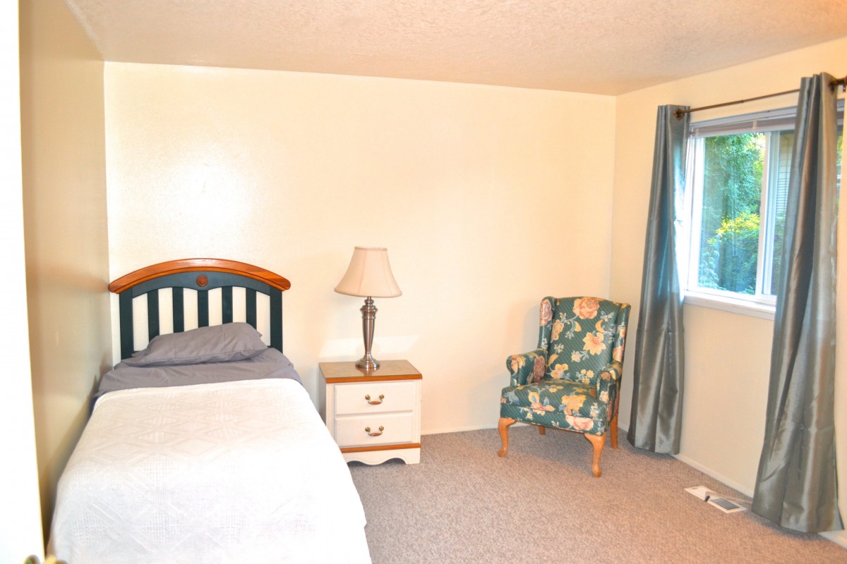 A simple bedroom with a bed, nightstand, lamp, and a patterned armchair near the window with curtains.