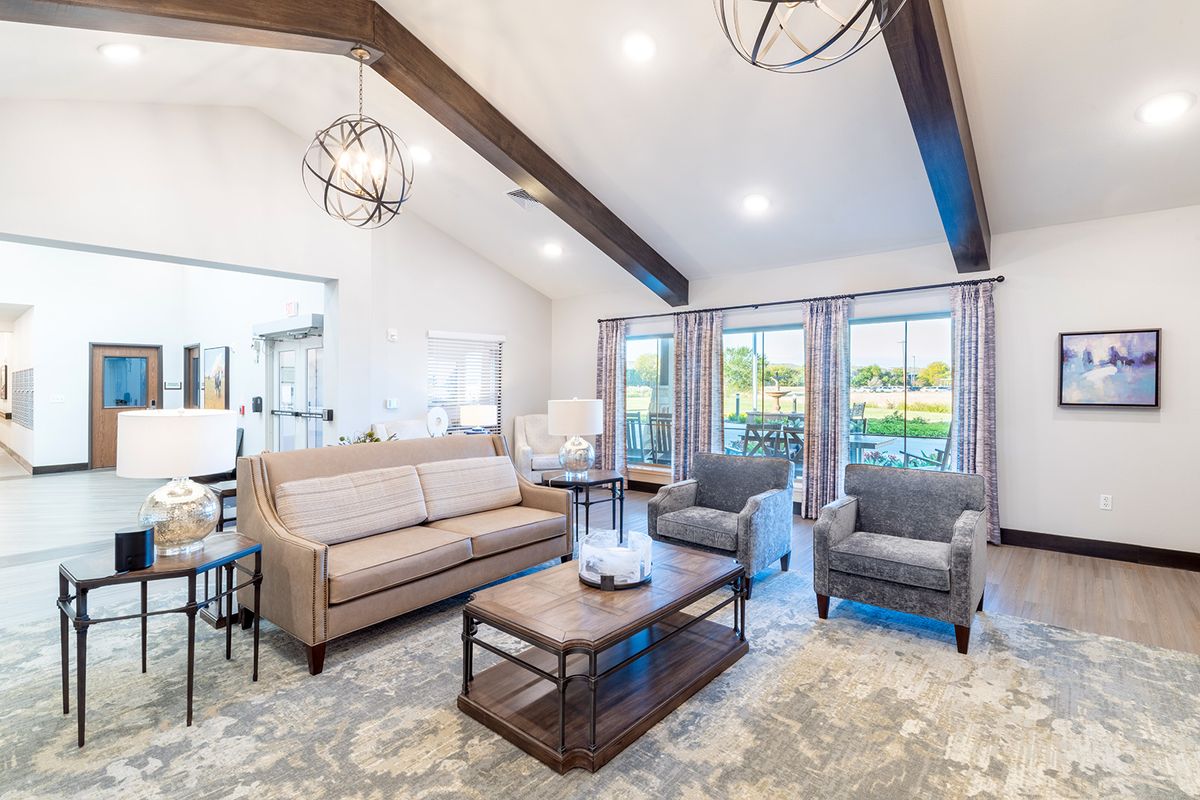 A modern living room featuring a comfortable sofa, two armchairs, and a wooden coffee table with decorative items. The space is brightened by large windows and accented with ceiling beams and a stylish chandelier.