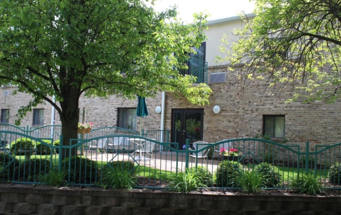 A view of a stone building with a patio area, surrounded by green trees and shrubs.