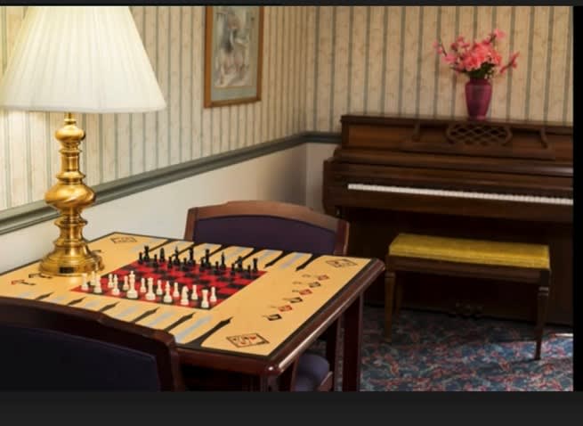 A cozy room featuring a chessboard on a table, with a lamp and piano in the background.