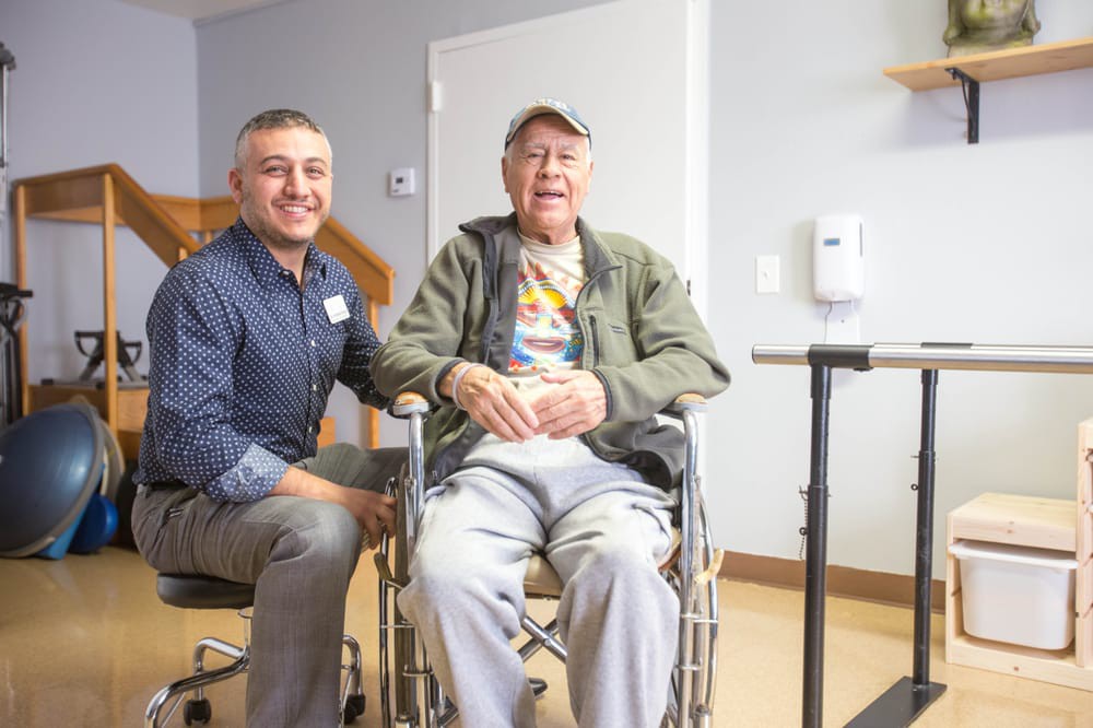 An elderly man in a wheelchair is accompanied by a caregiver inside a healthcare facility.