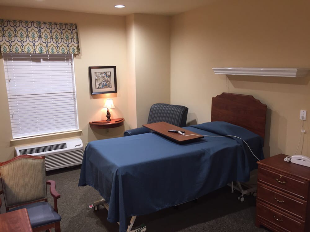 A neatly organized room featuring a hospital bed, chairs, and a lamp by the window.