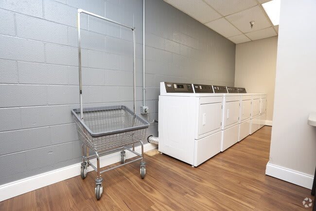 An empty laundromat with washing machines and a laundry cart against a gray wall.