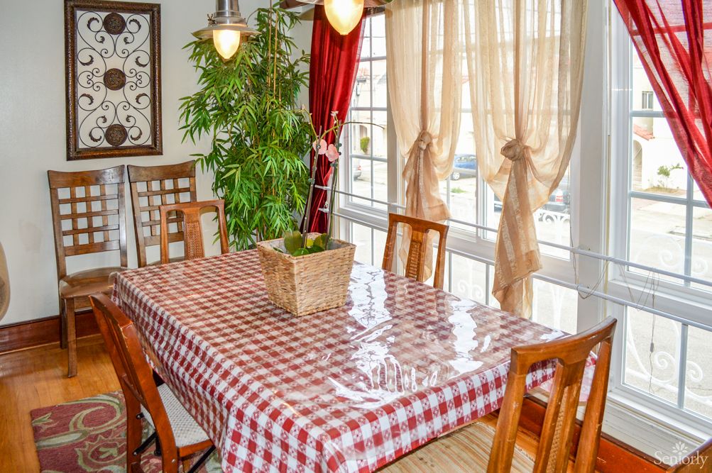 A cozy dining room featuring a checkered tablecloth, wooden chairs, and ample natural light from large windows.