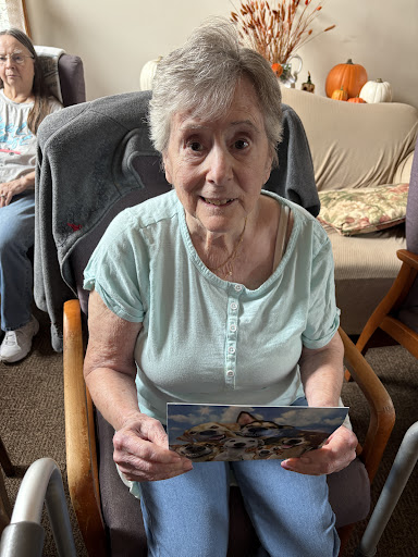 An elderly woman smiles while holding a postcard in her hands, seated comfortably indoors.