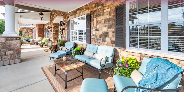 A cozy patio with cushioned chairs and a coffee table against a brick wall, accented by plants and large windows.