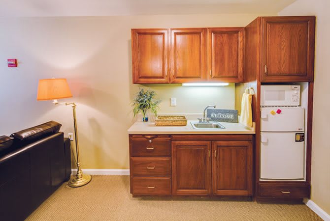 A small kitchen area with wooden cabinets, a sink, and a countertop next to a cozy lamp.