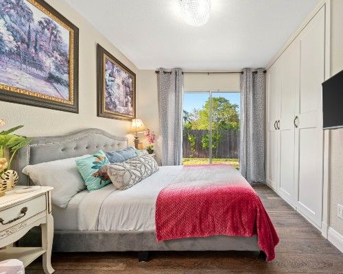 A cozy bedroom with a neatly made bed adorned with colorful pillows and a red blanket, complemented by artwork on the walls and a window with curtains letting in natural light.