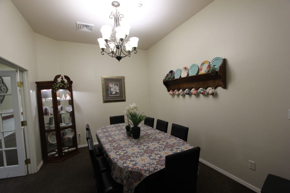 A cozy dining room featuring a table with a floral tablecloth, surrounded by chairs, and illuminated by a chandelier. Decorative plates adorn the wall, and a vase with flowers sits in the center of the table.