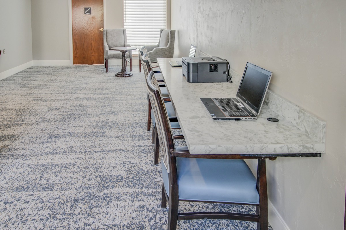 A quiet office space featuring a laptop and printer on a long table, with comfortable chairs and carpeted flooring.