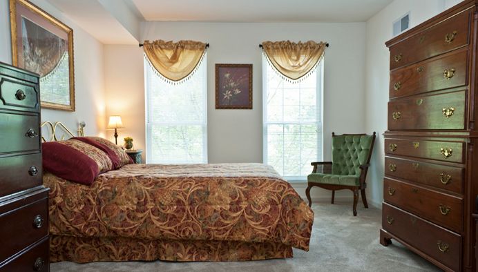 A cozy bedroom with a large bed, green armchair, and decorative curtains framing two tall windows.