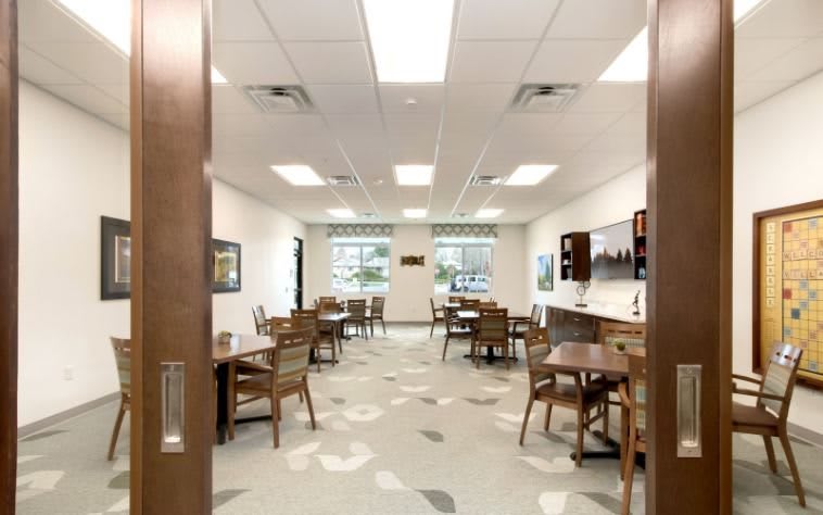 An interior view of a bright dining room with multiple tables and chairs, featuring large windows and ceiling lights.