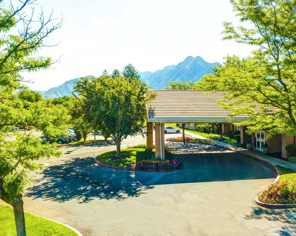 A hotel entrance with a driveway surrounded by trees and mountains in the background on a sunny day.