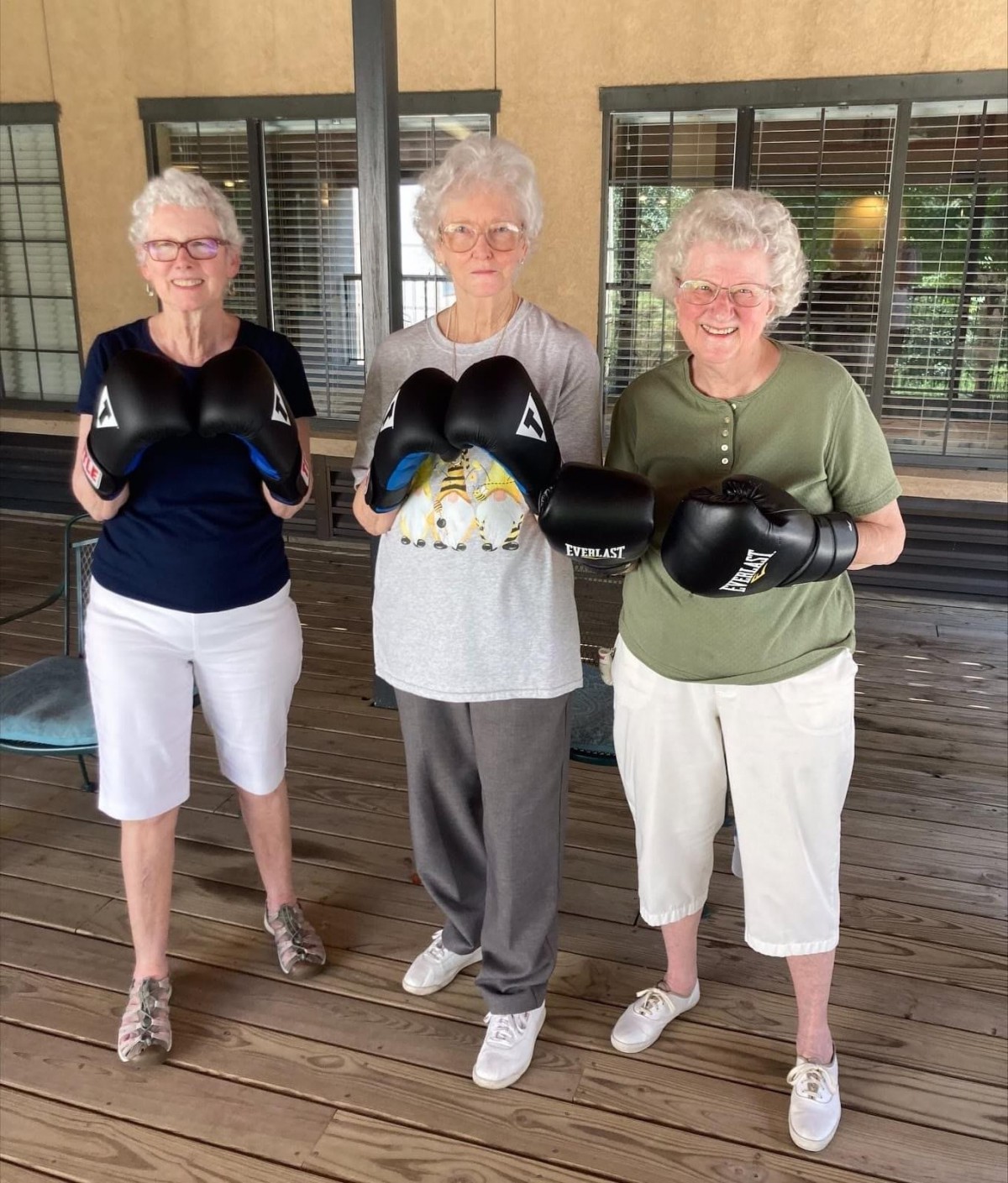 Three elderly women wearing boxing gloves stand together on a wooden deck.