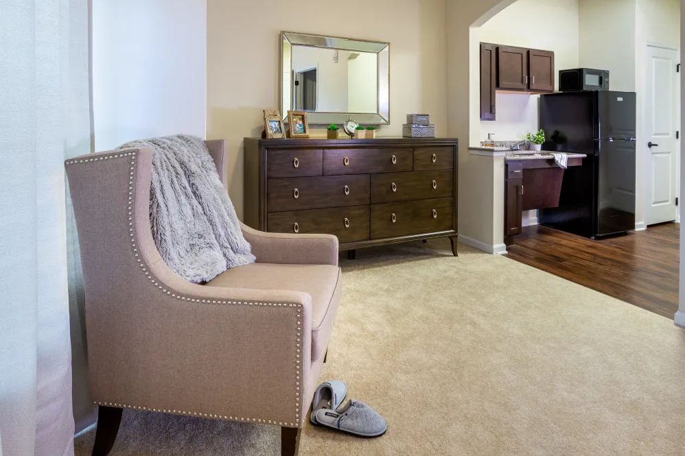 A cozy living room with a beige chair, dresser, and mirror, featuring comfortable slippers on the carpet.
