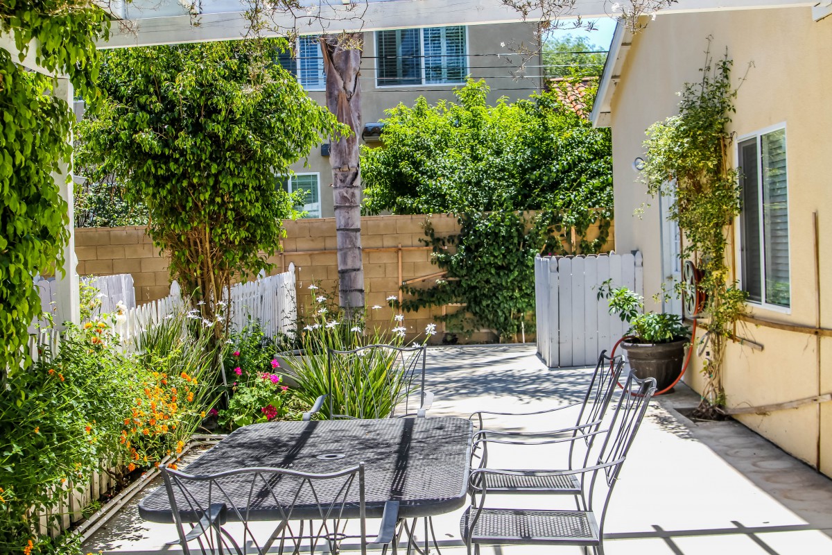 A sunny backyard patio with outdoor furniture surrounded by lush greenery and a wooden fence.
