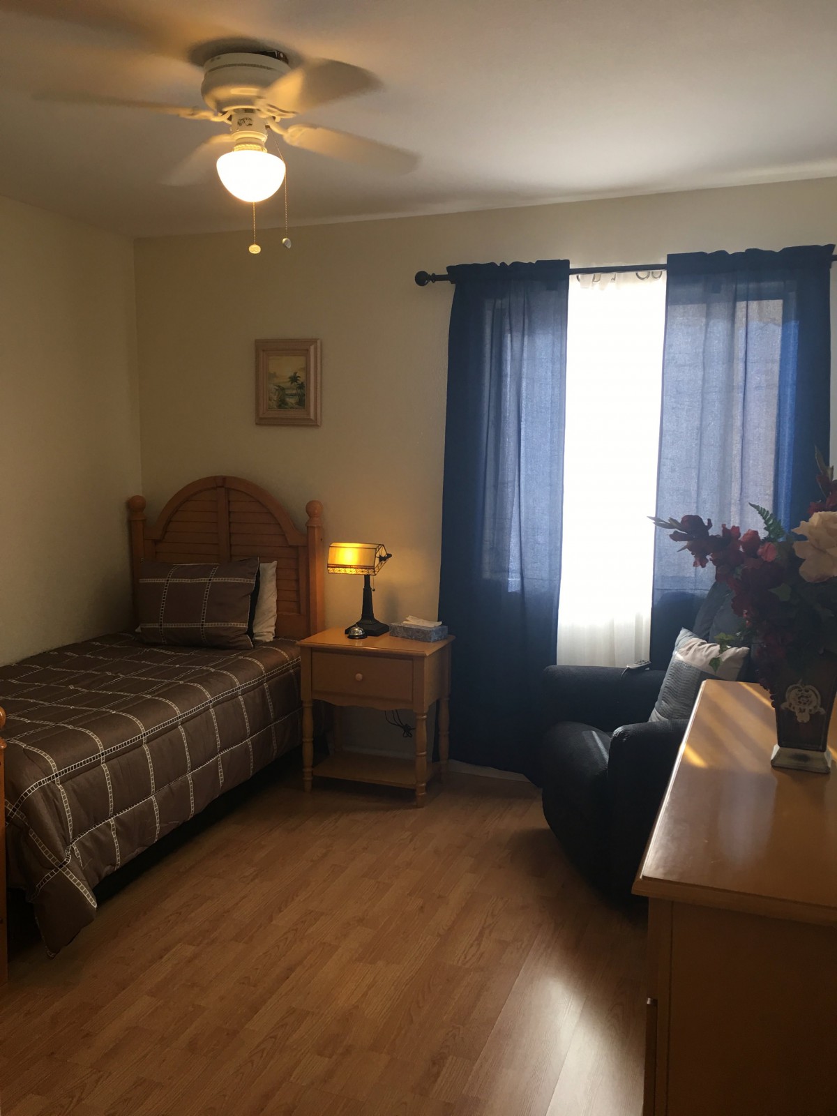A cozy bedroom with a single bed, nightstand, and armchair next to a window with blue curtains.
