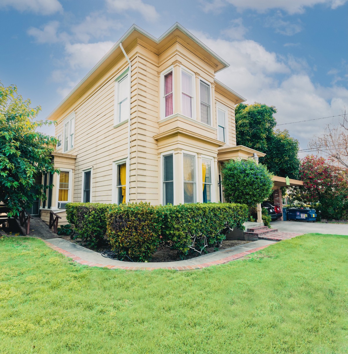 A charming two-story yellow house surrounded by a well-maintained garden and lush green lawn under a partly cloudy sky.