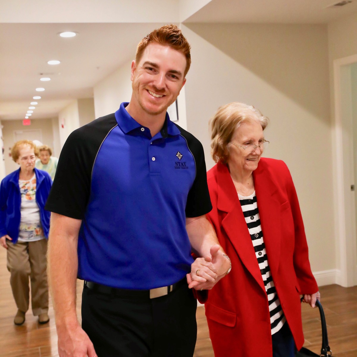 A caregiver walking with an elderly woman down a hallway, both smiling.