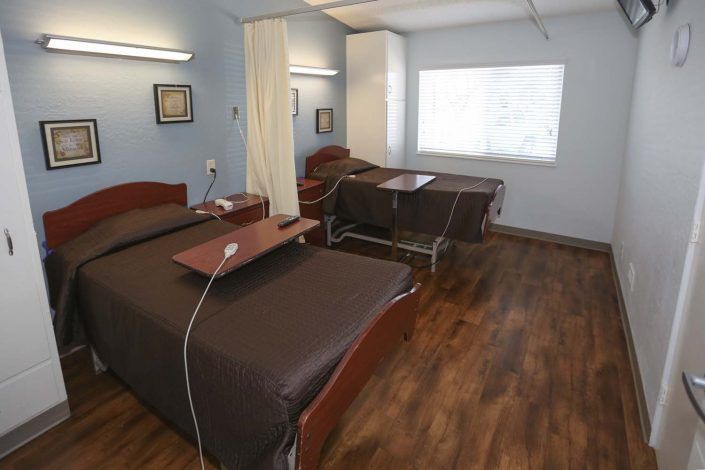 A hospital room with two patient beds, separated by a curtain, featuring wooden flooring and medical equipment.