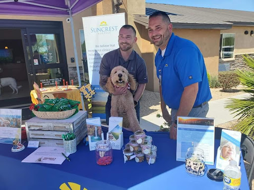 Two smiling individuals posing with a dog at an outdoor event booth.