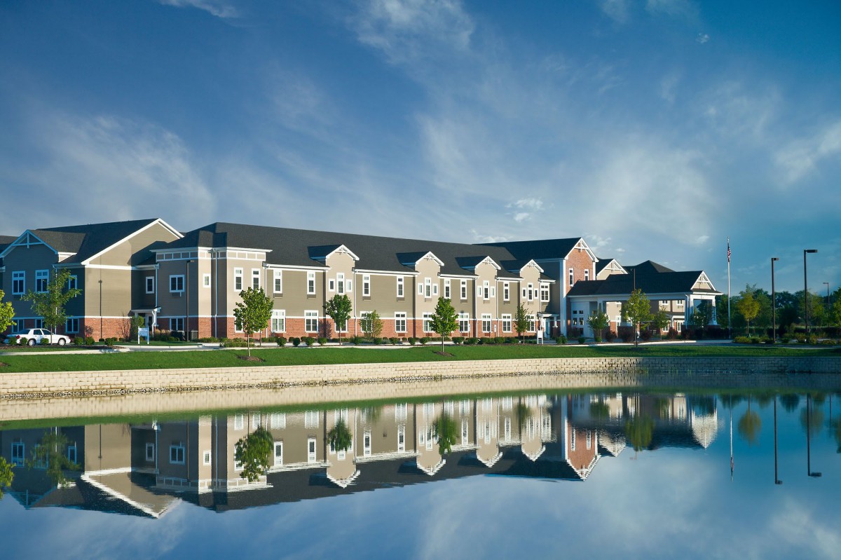 A residential building reflected in a calm lake with a clear blue sky and scattered clouds.