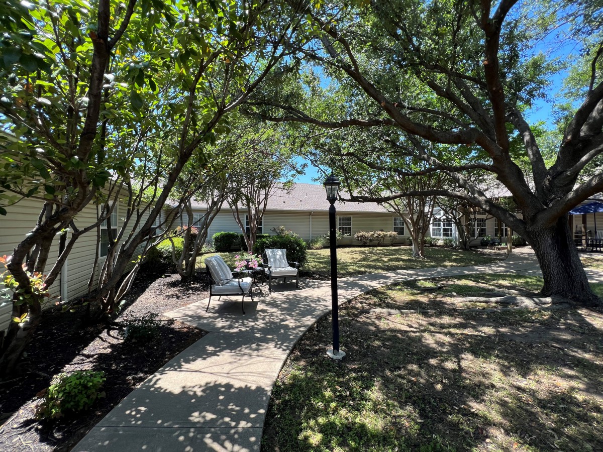 A tranquil outdoor garden with chairs under the shade of trees along a paved pathway.