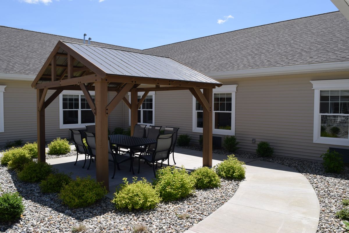 A sunny patio area features a wooden pergola with outdoor furniture, surrounded by landscaping.