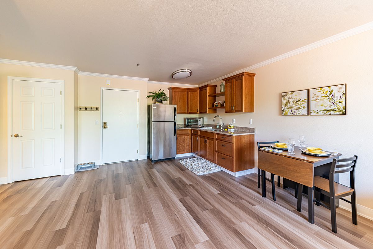 A cozy apartment kitchen with wooden cabinets and flooring, featuring a refrigerator and dining area.