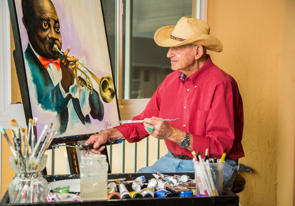 An artist wearing a cowboy hat works on a painting of a musician playing the trumpet.