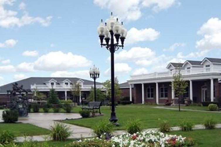 A scenic view of a garden with a vintage lamppost, benches, and surrounding buildings under a partly cloudy sky.