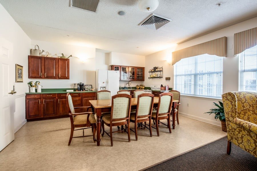 A spacious kitchen and dining area featuring a large wooden dining table with upholstered chairs, surrounded by cabinets and a window letting in natural light.