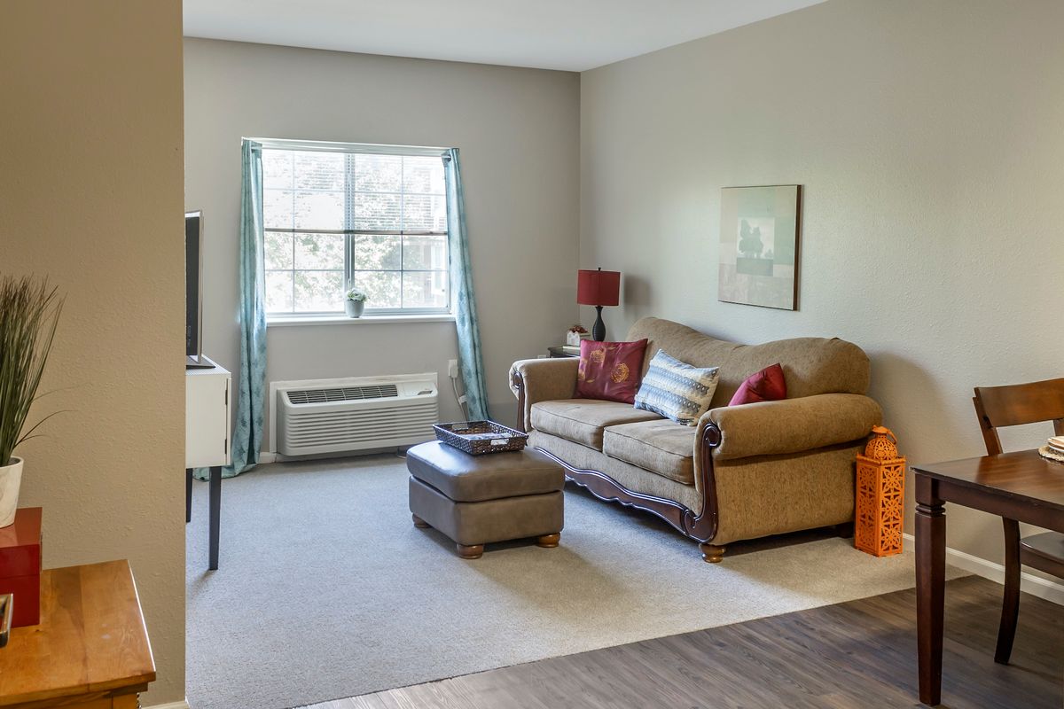 A cozy living room with a plush sofa and ottoman, decorated with cushions and artwork, featuring a large window that brightens the space.