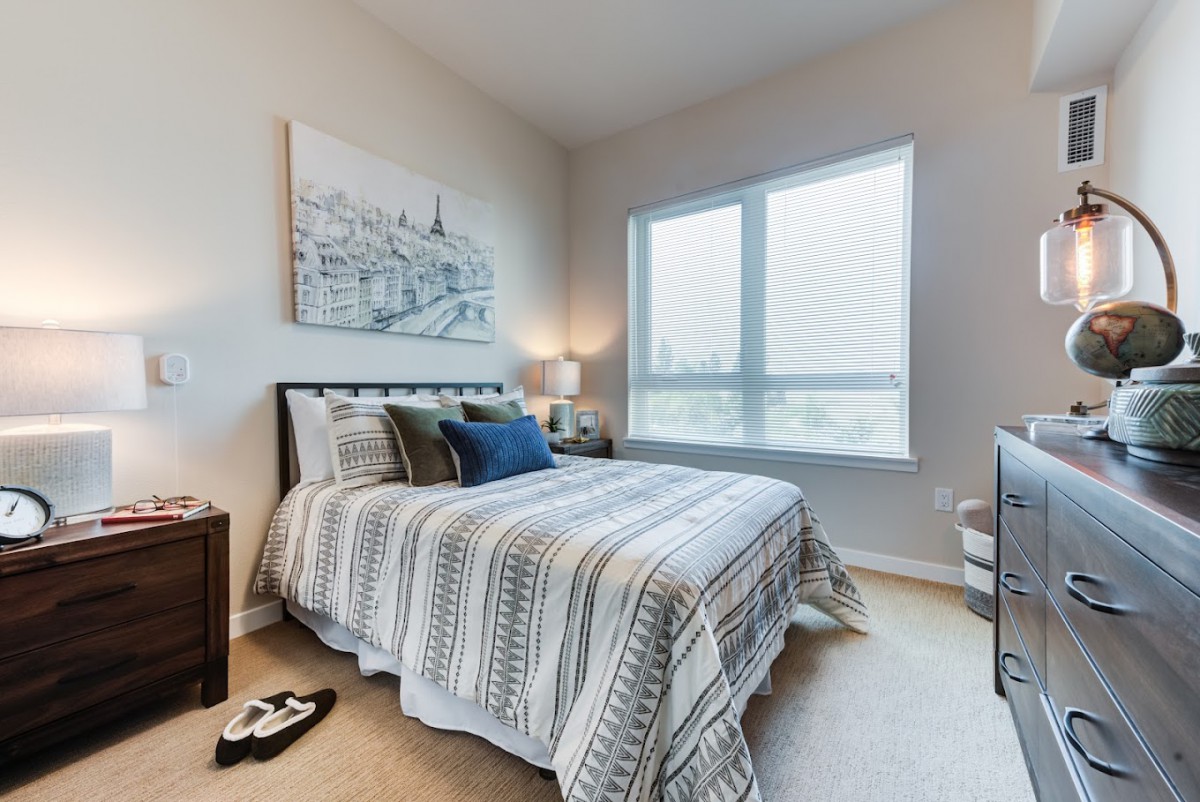 A cozy bedroom with a neatly made bed, artwork above the headboard, and natural light streaming through the window.