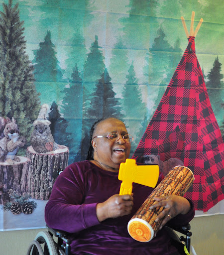 A cheerful woman in a wheelchair holds a toy axe and a drum, set against a vibrant forest-themed mural.