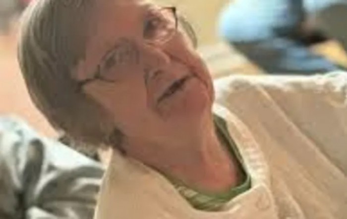 An elderly woman wearing glasses and a sweater, sitting indoors.