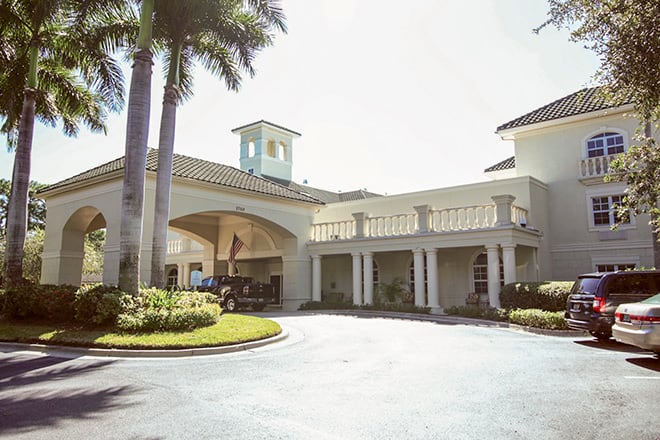 A sunny view of a building entrance with palm trees lining the driveway.