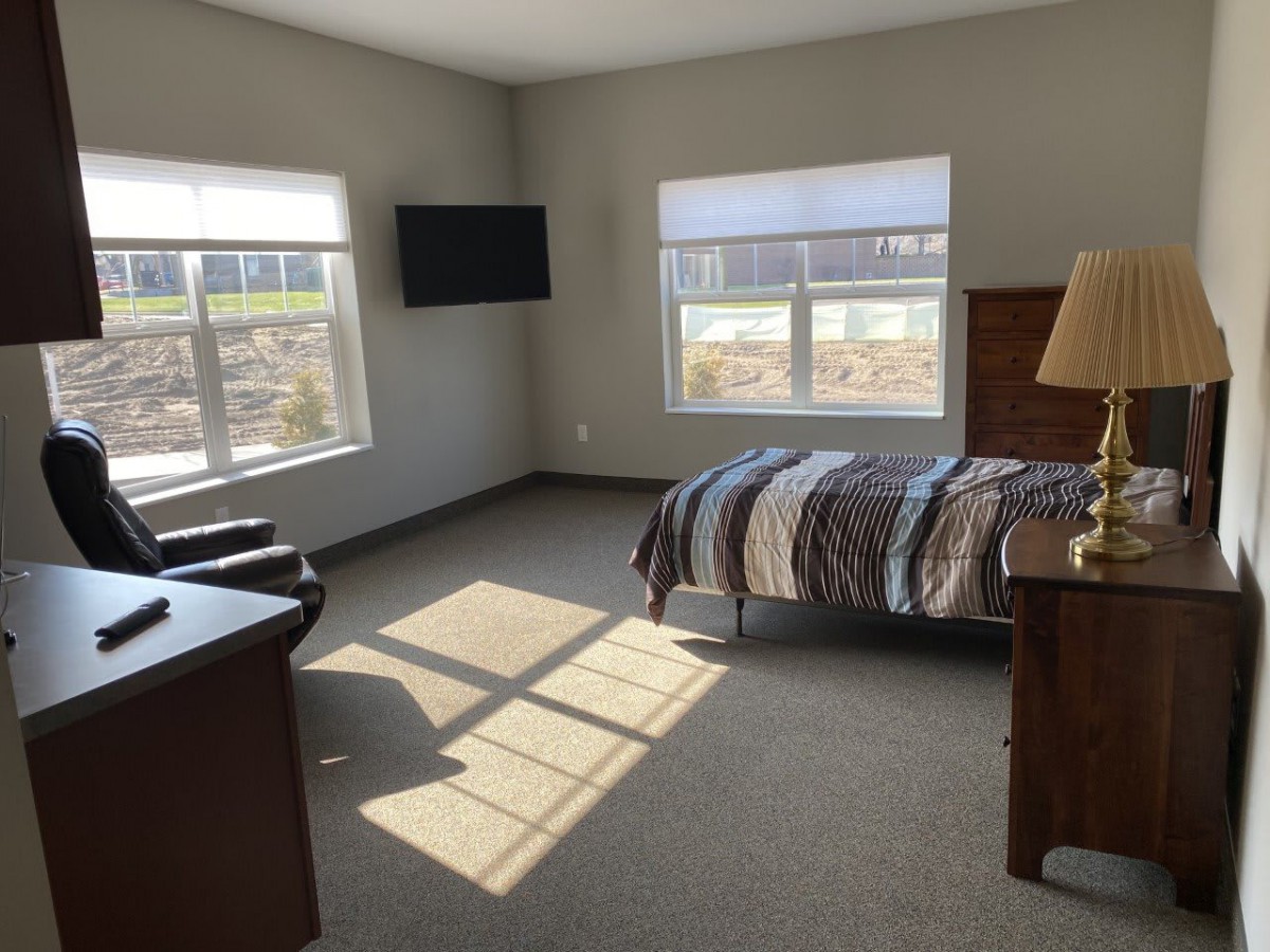 A well-lit bedroom with a single bed, a television mounted on the wall, and two large windows allowing natural sunlight to fill the space.