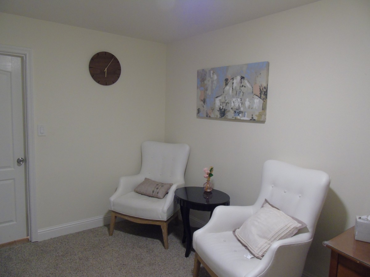 A cozy living room corner with two white chairs, a small table, wall art, and a clock.