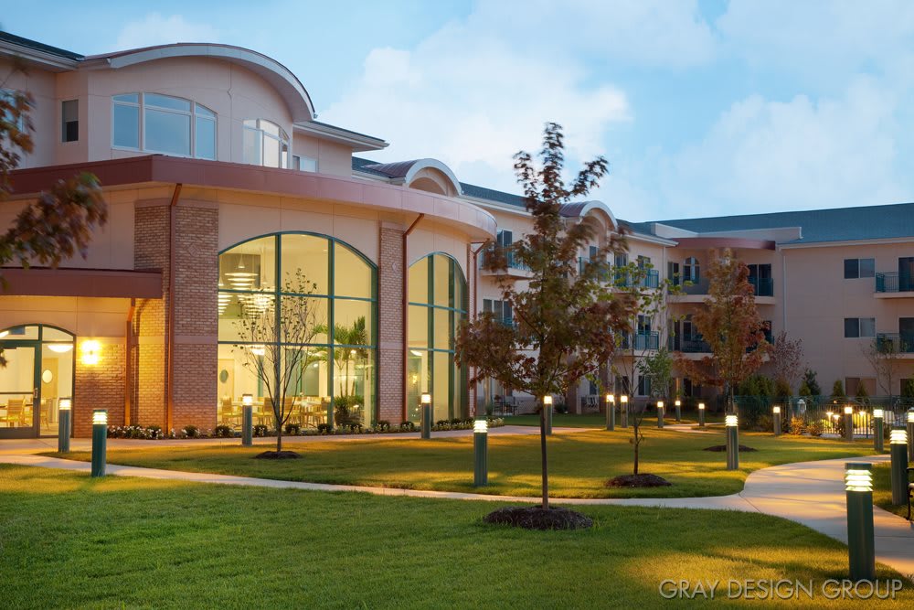 A modern building with large windows, surrounded by a well-lit garden and trees during the evening.