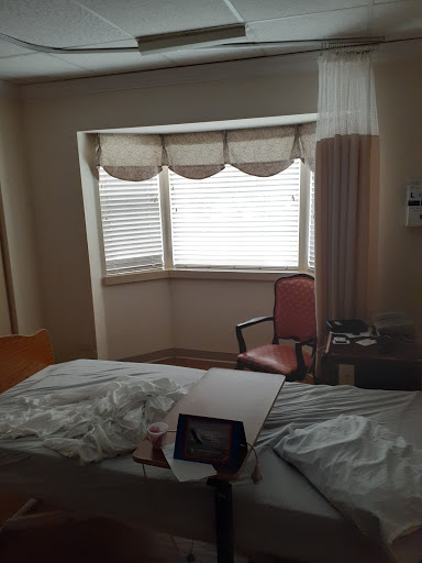 A view of a hospital room featuring a bed, a window with curtains, and a chair.